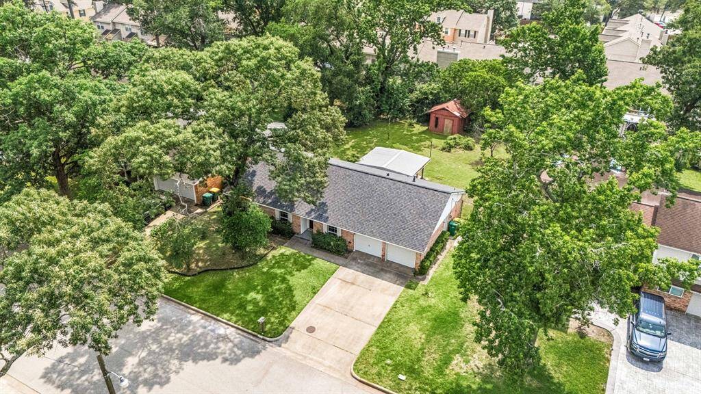This aerial view showcases a charming single-story home with a well-maintained roof and driveway. The property is surrounded by lush greenery and mature trees, offering privacy and shade. There's a spacious backyard with a small shed, perfect for storage or a workshop. Situated in a quiet neighborhood, it provides a serene setting ideal for family living.