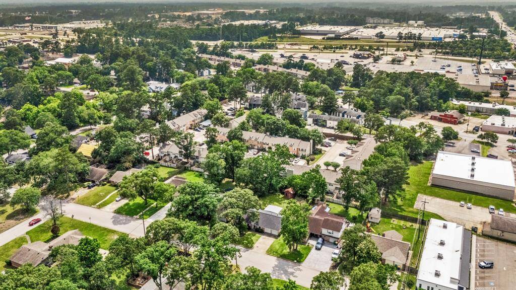 Aerial view of a suburban neighborhood with tree-lined streets and a mix of single-family homes and townhouses. The area is lush with greenery and offers proximity to commercial buildings and shopping centers in the background, providing convenience and a residential feel.