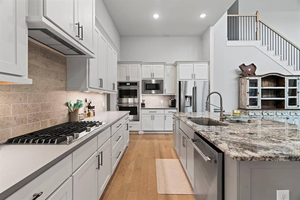 Chef's kitchen perfection. Crisp white cabinetry, granite counters, and a spacious island offer beauty and function in this gourmet-ready space. Cabinetry, stainless steel appliances, granite countertops, and a gas cooktop. There's ample storage and a high ceiling, with a view of a staircase leading to the upper level.