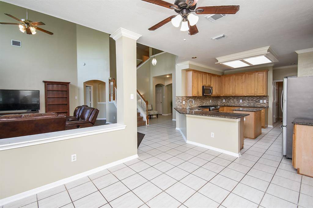 This modern kitchen features granite countertops, stainless steel appliances, and ample wooden cabinetry. The open layout connects to a bright living area with large windows and high ceilings, offering a spacious and inviting atmosphere.