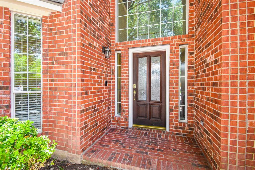 This photo showcases a welcoming brick entrance with a wooden front door, flanked by tall windows. The entryway is bright with abundant natural light from large, overhead windows, providing a spacious and inviting feel.