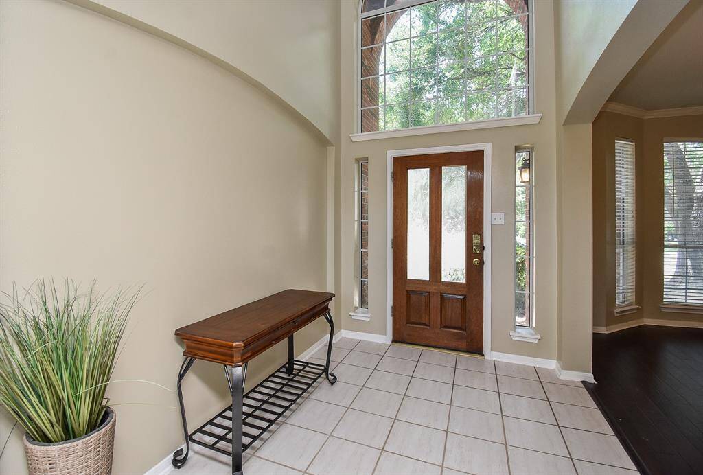 This entryway features a high ceiling and large windows for abundant natural light. It includes a wooden front door with frosted glass panes, tiled flooring offering a welcoming and spacious feel.