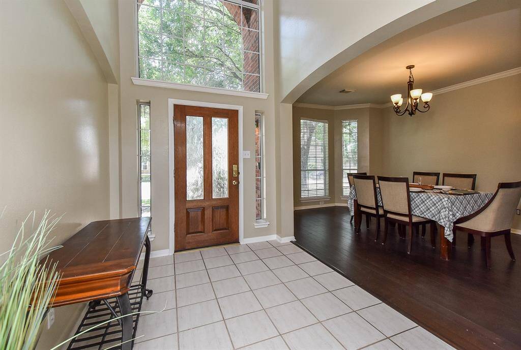 This photo showcases a bright entryway with tiled flooring and a wooden front door featuring decorative glass panels. Adjacent is a dining area with dark wood flooring, a stylish chandelier and neutral paint throughout.