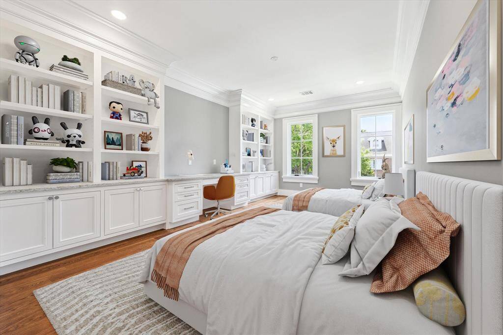 This bright and spacious bedroom features two twin beds, built-in shelving with lots of storage, and a desk area. The large windows provide plenty of natural light, complementing the elegant crown molding and warm wood flooring. There is also a large walk-in closet and an en-suite bath.  This photo has been virtually staged.