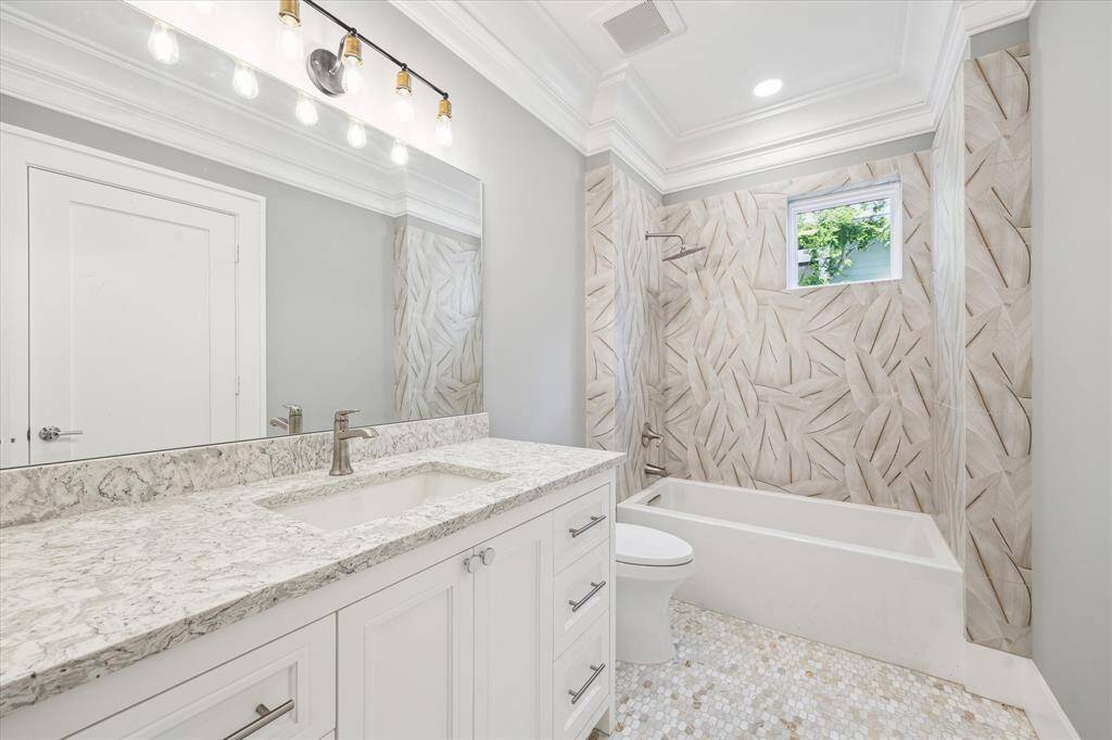 This secondary bathroom features a modern design with a large vanity and marble countertop, a sleek bathtub with decorative tiled walls, and a bright atmosphere enhanced by recessed lighting and a small window.