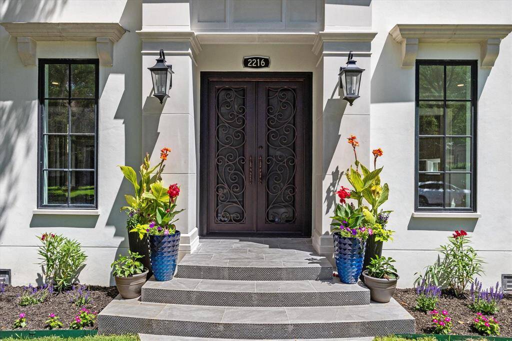 This elegant front entrance with dark iron ornate doors are framed by large widows and stylish lanterns. The area is beautifully landscaped with colorful potted plants, creating a welcoming and sophisticated curb appeal.