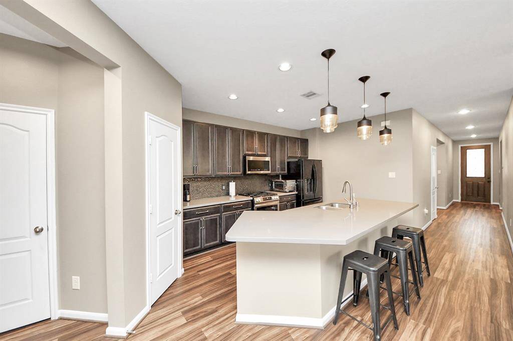 This spacious kitchen features highceilings, dark stained wood cabinets, SS appliances,modern tile backsplash, recessedlighting, and kitchen islandwith double sink.