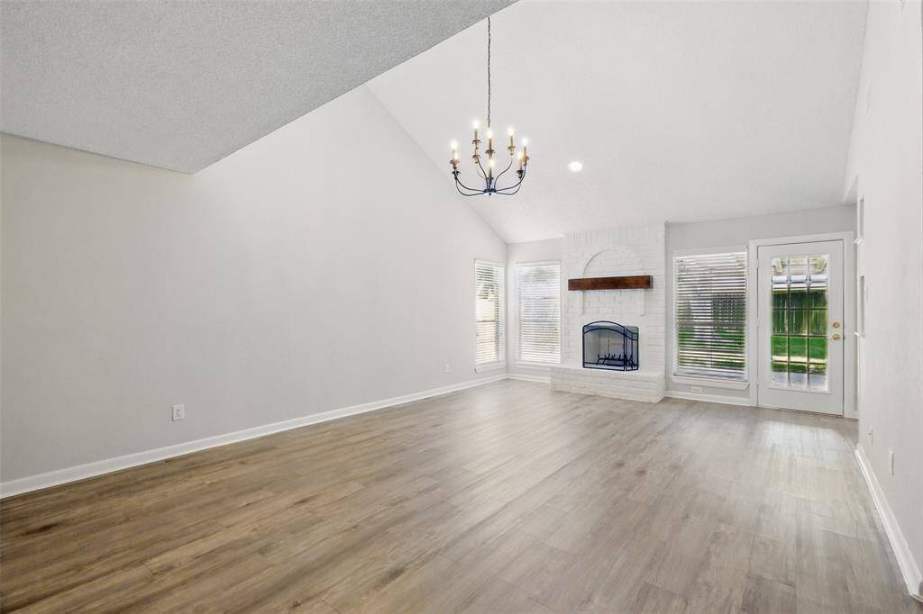 Bright and spacious living room with vaulted ceilings, a modern chandelier, and vinyl flooring. Features a white brick fireplace and large windows for natural light, plus a glass door leading to the backyard.