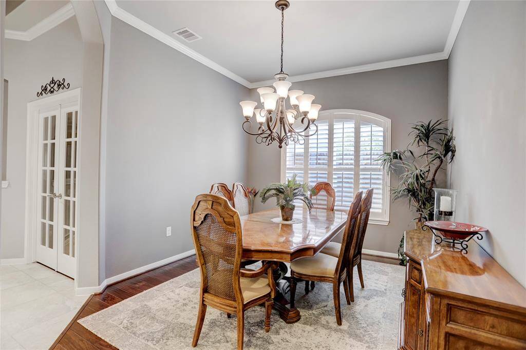 An elegant dining room features a chandelier and large windows with plantation shutters, set on a neutral color palette with crown molding.