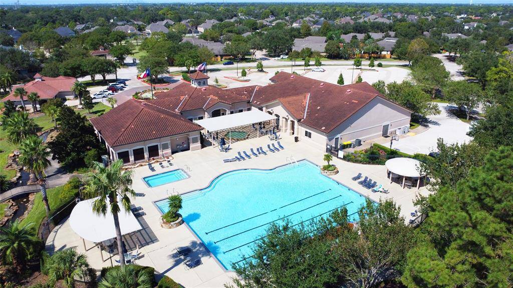 This aerial photo shows a spacious community clubhouse with a large outdoor swimming pool surrounded by lounge chairs, lush greenery and ample parking. Not shown, is the neighborhood's large, stocked pond with concrete walking trail encircling.