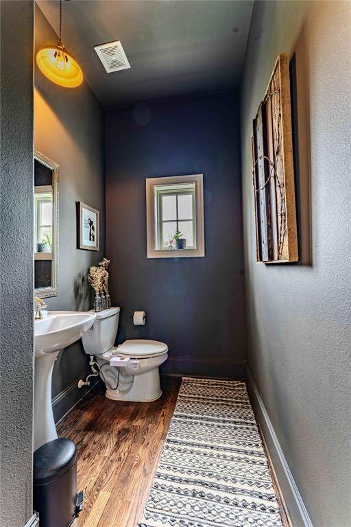 This is a cozy half-bathroom with a modern design. It features a pedestal sink, a toilet, and a small window that brings in natural light. The dark walls contrast with the wood flooring, bringing the same sophisticated luxury into this perfectly located half bath.