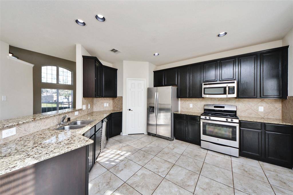 This modern kitchen features dark cabinetry, granite countertops, and stainless steel appliances, including a gas stove and double-door refrigerator. The space is bright with natural light and has a tiled floor, offering both functionality and style.