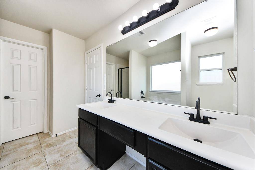 This bathroom features a spacious double vanity with modern black fixtures, a large mirror with ample lighting, tiled flooring, and a combination of natural and overhead light, creating a bright and functional space.