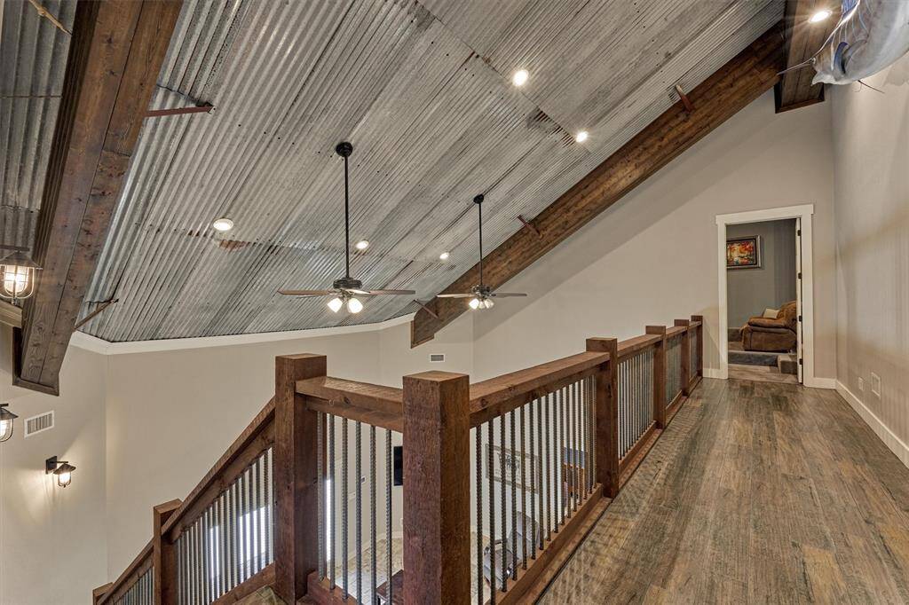 The second floor showcases a perfect blend of rustic and modern design, featuring a tin-style ceiling and exposed wooden beams. The balcony area, which overlooks the main living space, leads to the media room and highlights a custom cedar wood railing with rebar spindles—beautifully tying together the home’s unique architectural elements.
