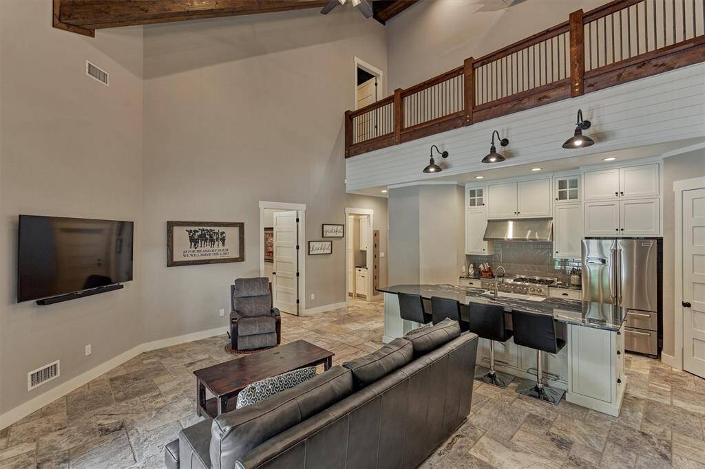 This view highlights the second-floor catwalk that leads to the media room, with striking wood beams accenting the ceiling and creating a warm, rustic touch. The open-concept kitchen is visible below, while doorways beyond lead to a private bedroom and full bath.