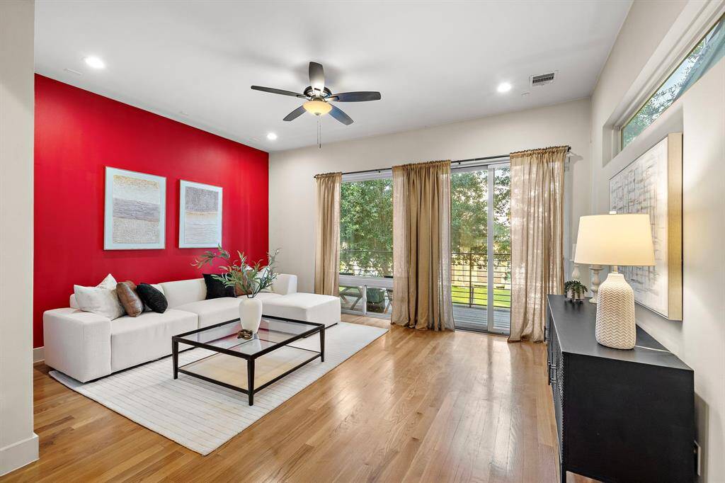 Contemporary living room features a bold red accent wall, sleek ceiling fan, and floor-to-ceiling windows that flood the space with natural light. Enjoy direct access to a private balcony and an airy, open layout with modern hardwood floors.