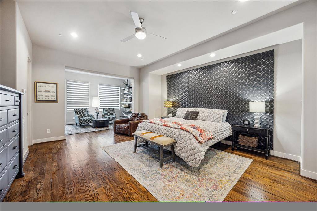 The spacious primary bedroom serves as a serene retreat, featuring soft natural light, a removable textured accent wall that adds modern character, and warm hardwood floors throughout. A cozy seating nook with a built-in bookshelf offers an inviting space to read, unwind, or simply enjoy the tranquil atmosphere.