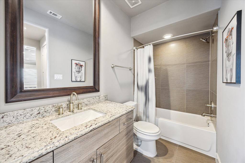 This stylish en suite bath offers modern functionality with a granite countertop, sleek fixtures, and a spacious tub-shower combination framed by contemporary tile. A large mirror enhances the sense of space, while the neutral color palette adds a clean, sophisticated finish. With a secondary entrance from the hallway, it provides both privacy and convenience for guests.