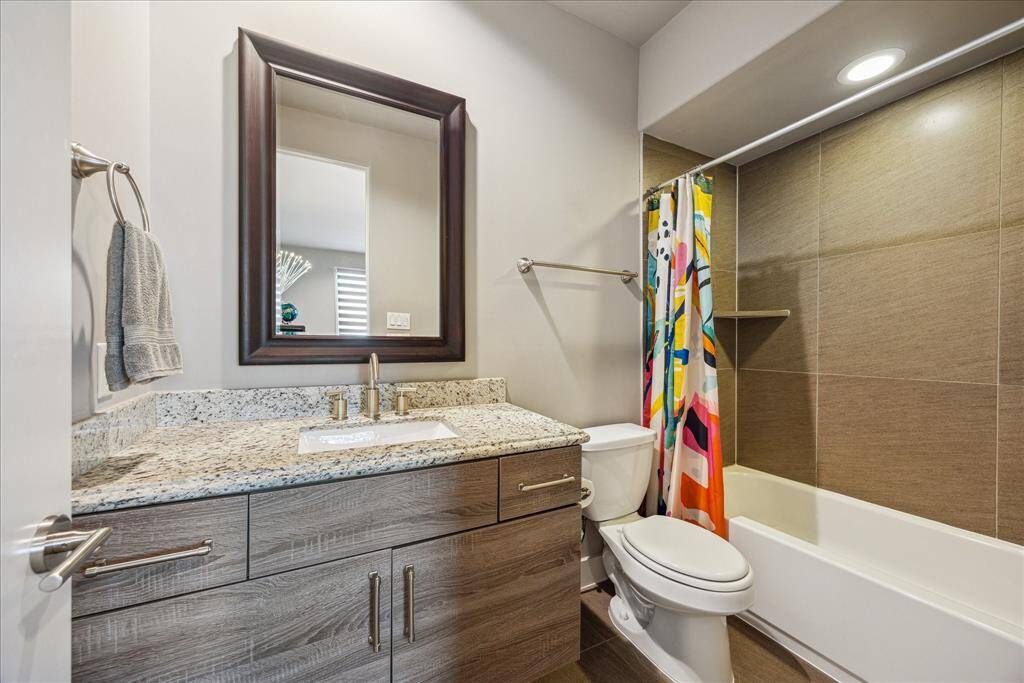 This en suite bath offers modern style and everyday convenience with granite countertops, sleek wood cabinetry, a large mirror, and a tub-shower combo surrounded by neutral tile.