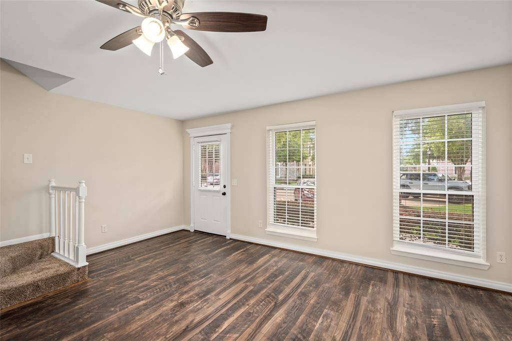 Inviting main living area featuring rich wood-style flooring, abundant natural light from three tall windows, and a neutral color palette. The space offers a seamless flow from the entryway and includes easy access to the staircase for upstairs living.