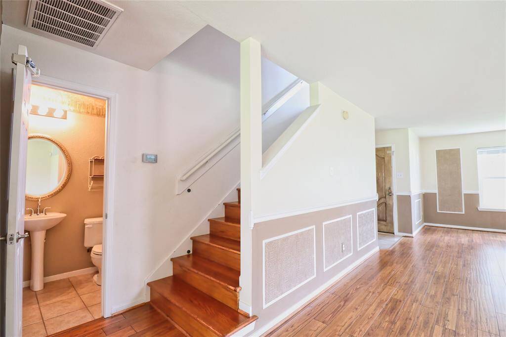 This photo showcases a bright, open area with hardwood floors and a staircase leading to the upper level. A small bathroom is visible on the left with a pedestal sink and tiled floor. The room has neutral walls, decorative paneling, and ample natural light.