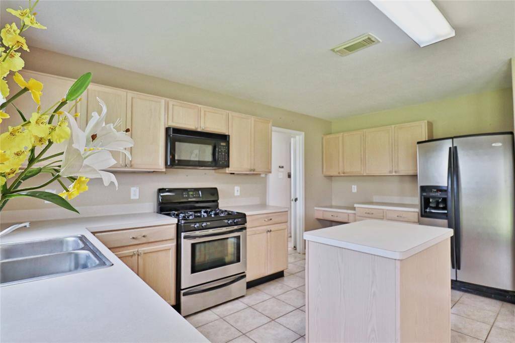 The kitchen features light wood cabinets, a central island, stainless steel appliances, and a gas stove. The space is bright and airy with tile flooring .