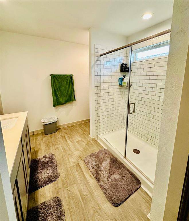 Bright bathroom with wood-style flooring, featuring a spacious glass-enclosed shower with subway tiles. Includes modern vanity and ample lighting.