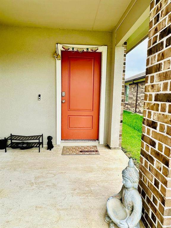 Front entryway featuring a bold red door, a welcoming mat, and decorative elements like a small bench and a serene statue. The area is framed by brickwork and opens to a lush green lawn, creating a warm, inviting atmosphere.