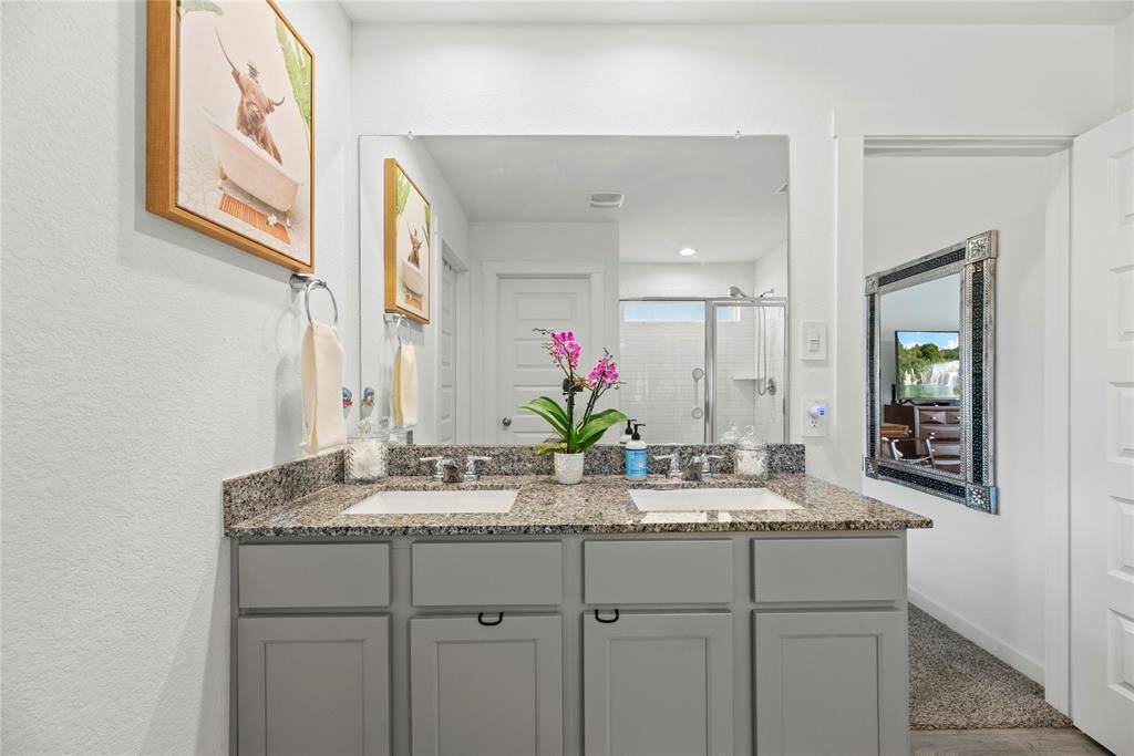 The primary bathroom vanity is designed with two square sinks, each equipped with stylish nickel faucets. The gray cabinets offer a modern, sleek look, while the light natural wood-colored LVP flooring adds warmth and a touch of nature to the space. This combination of finishes creates a sophisticated yet inviting atmosphere, balancing contemporary design with a touch of earthy charm. The polished nickel details complement the overall aesthetic, making the bathroom both functional and visually appealing.