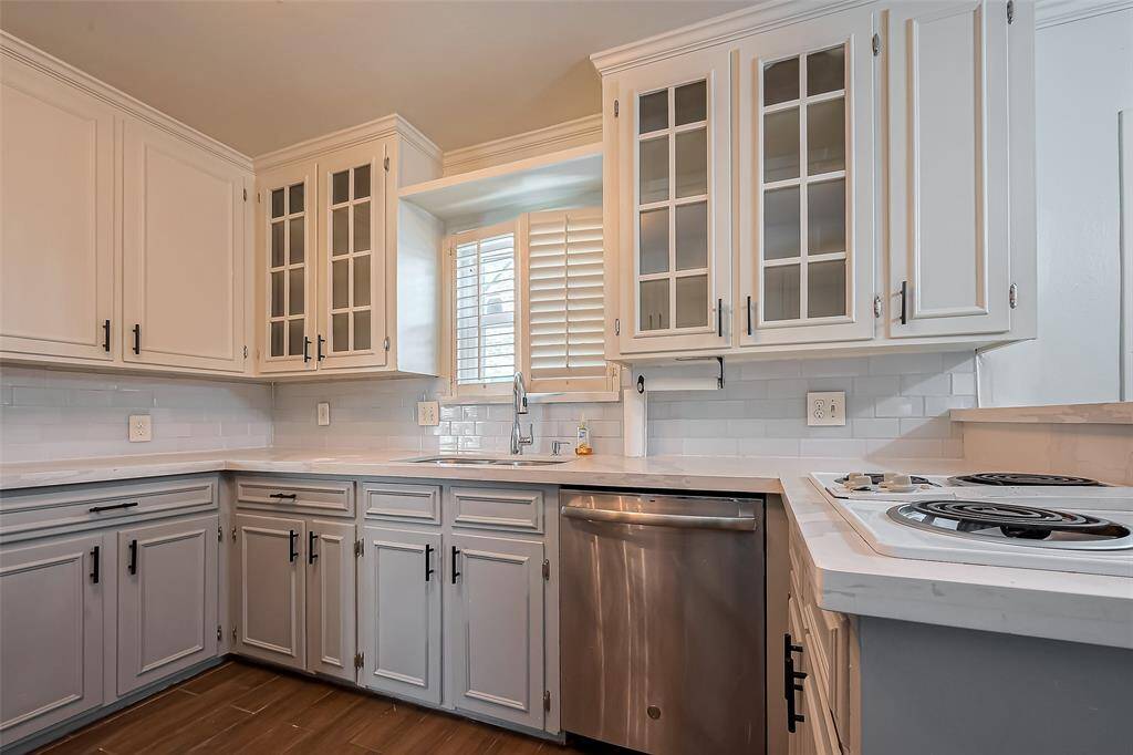 This kitchen features modern white cabinetry with glass-front accents, sleek countertops, and stainless steel appliances. A window with shutters provides natural light, complemented by a neutral backsplash and electric cooktop.