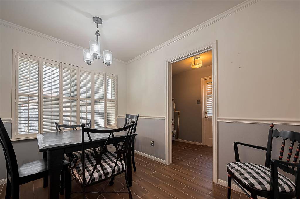 This dining area features a cozy, well-lit space with a modern light fixture, large windows with shutters, and a stylish black dining set. The room has a warm, inviting atmosphere with wood-look tile flooring and neutral wall tones.