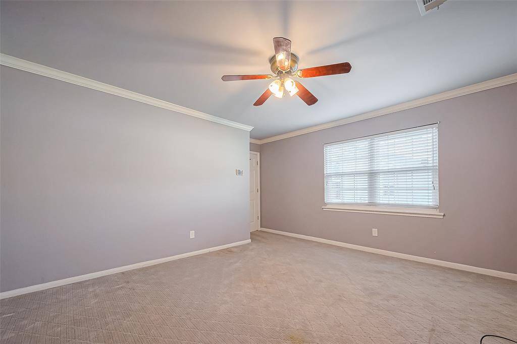 This photo shows a spacious, carpeted bedroom with soft gray walls, featuring a ceiling fan with lights. It has a large window with blinds, providing ample natural light.