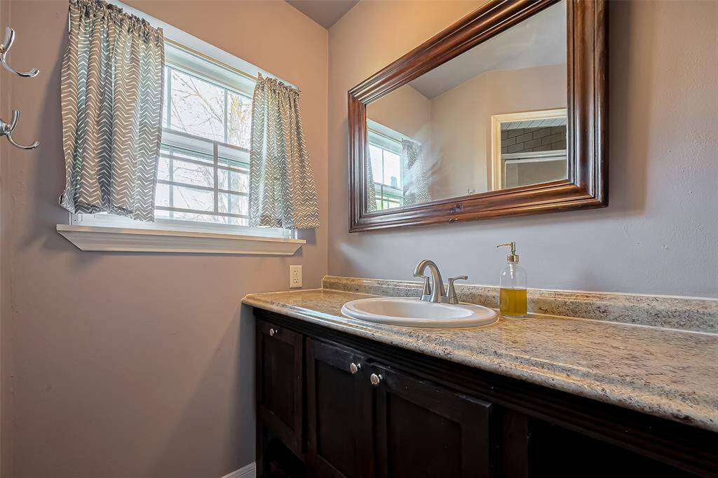 This is a bright bathroom with a granite countertop and single sink. It features a large mirror, under-sink storage with dark cabinetry, and a window with patterned curtains that allow natural light in.