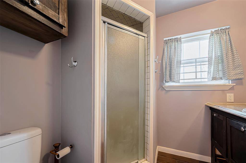 This bathroom features a compact design with a frosted glass shower, natural light from a window with patterned curtains, and a dark wood vanity for storage. Ideal for efficient use of space.