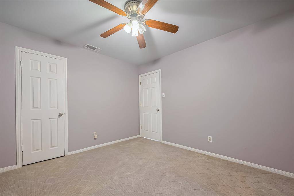 This room features soft gray walls, neutral carpeting, and a ceiling fan with wooden blades. It has two doors and a clean, spacious feel, ideal for a bedroom or office.