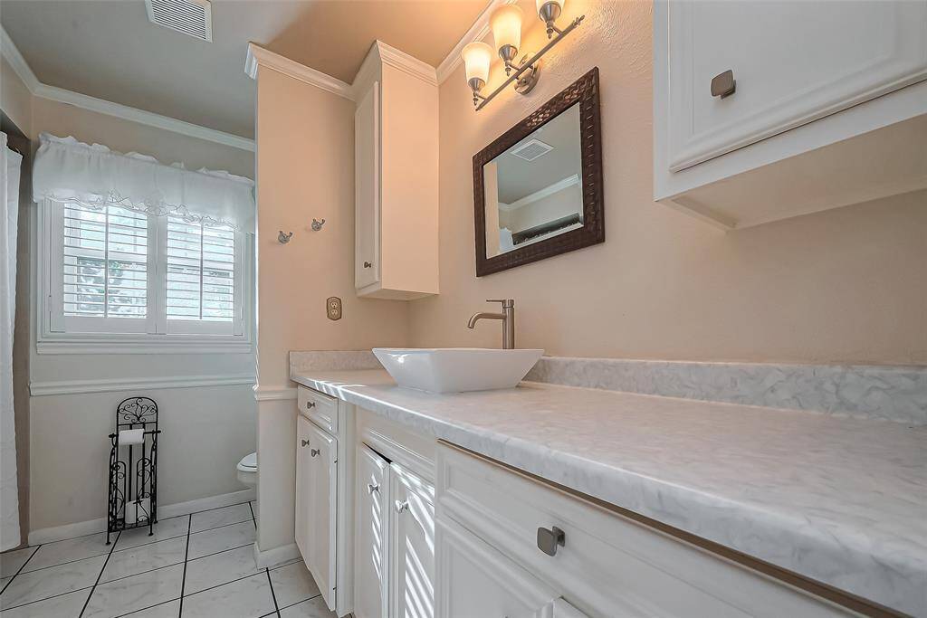 This room features soft gray walls with white trim, carpeted flooring, and a ceiling fan with wooden blades. A large window with shutters allows natural light to brighten the space, creating a cozy and inviting atmosphere.