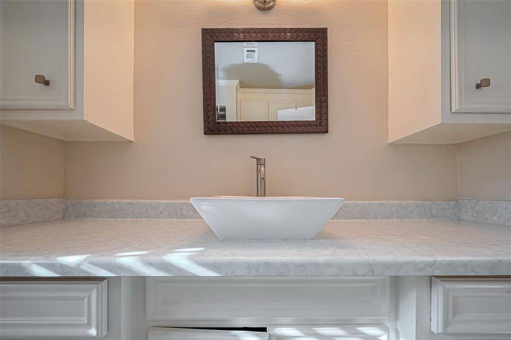 This bathroom features a sleek modern design with a vessel sink, marble countertop, and ample cabinet storage. Natural light filters through a window with shutters, complementing the neutral color palette.