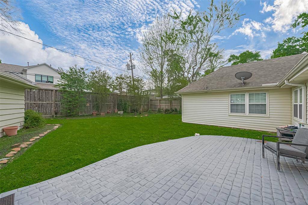 This backyard features a spacious grass lawn with a privacy fence and mature trees. There's a large, paved patio area perfect for outdoor seating or entertainment. The home's exterior is well-maintained, with a neutral color palette and large windows.