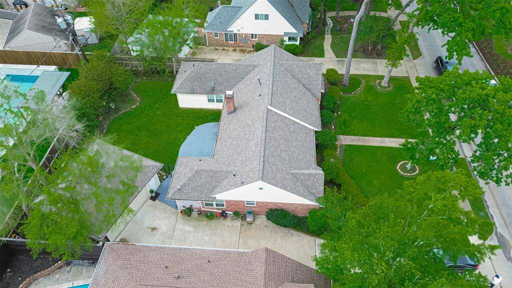 This aerial view shows a charming single-story home with a well-maintained roof and a spacious driveway. It's surrounded by lush greenery and mature trees, offering a welcoming front yard and a cozy backyard space. The neighborhood appears quiet and residential, perfect for those seeking a peaceful living environment.