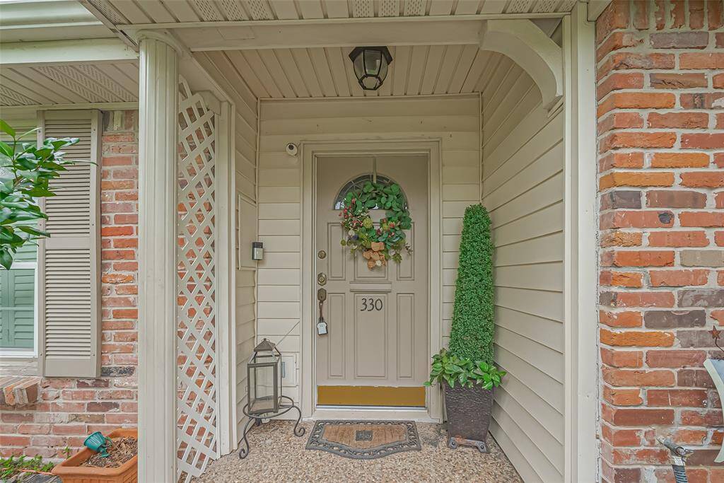 This inviting front entry features a charming door with a decorative wreath, nestled between red brick and light siding. A lantern and potted plants add a welcoming touch, complemented by a trellis and exterior light.