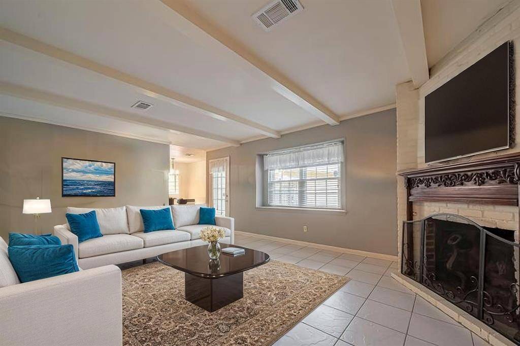 Virtual Staging - This cozy living room features a light color palette with a comfortable sectional sofa accented by blue pillows. A large window allows natural light to fill the space. The room includes a decorative fireplace with a mounted TV above, and a stylish coffee table on a patterned rug completes the inviting setup.