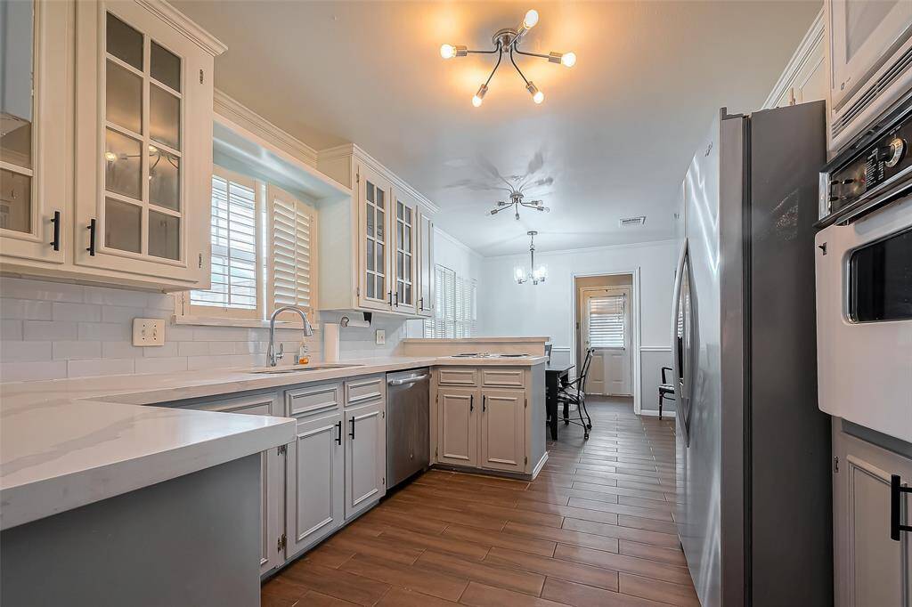This kitchen features sleek stainless steel appliances, white cabinetry with glass accents, and a stylish light fixture. The space is bright with ample natural light and has a modern, open layout with a dining area in view.