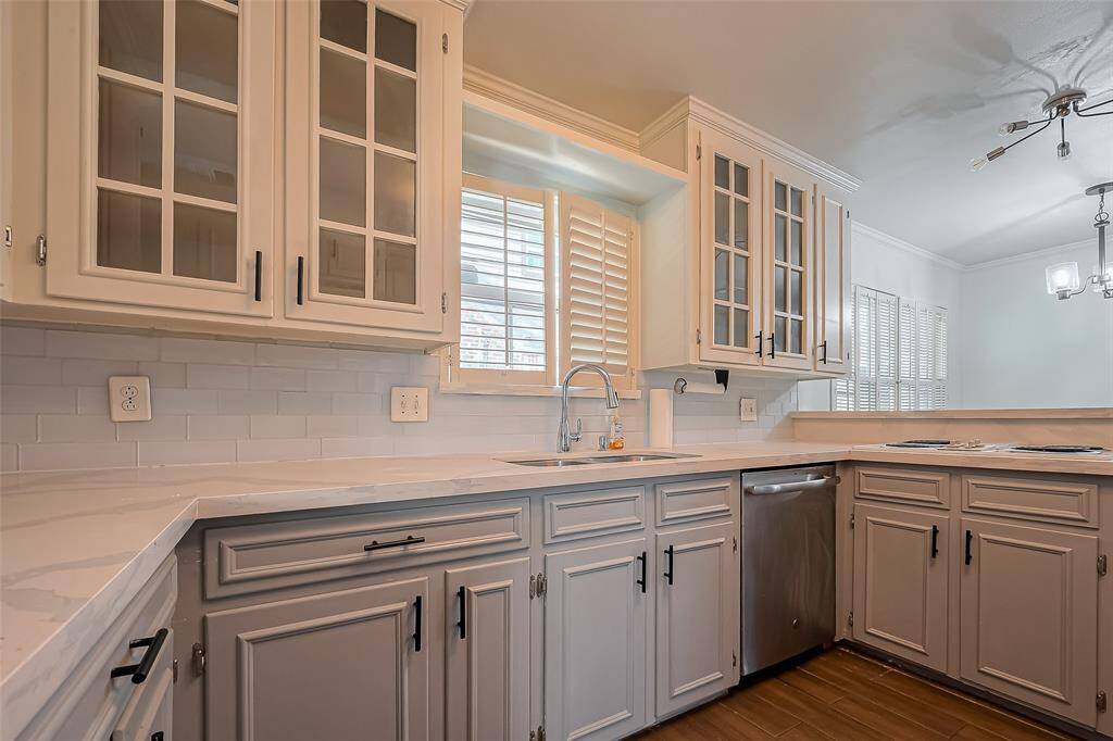 This kitchen features elegant white cabinetry with glass-paneled doors, a sleek undermount sink, and modern stainless steel appliances. The bright, airy space is enhanced by natural light from the window with shutters, complemented by a stylish light fixture above.
