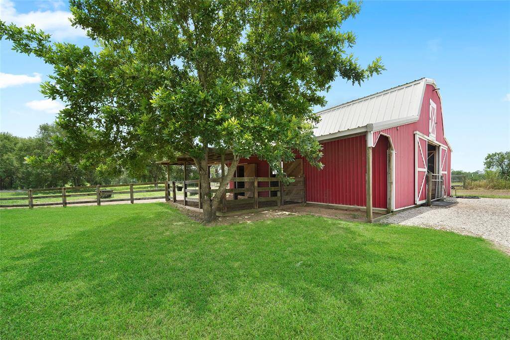 The side view of the barn, shaded by mature trees, adding to the peaceful country atmosphere. The wide driveway provides ample space for maneuvering vehicles and trailers.