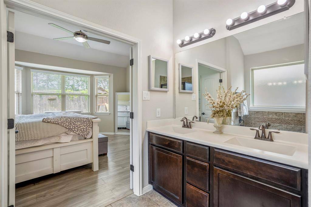 A close-up of the dual sinks reveals thoughtful details — the window’s soft light reflected in the mirror, and just to the left, the bay windows peek through the open double doors, tying the bathroom and bedroom together with warmth and natural light.