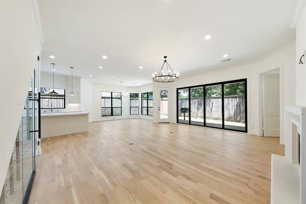 This spacious open-concept living area features light wood flooring, abundant natural light from large windows, and modern fixtures. It seamlessly connects to a bright kitchen with an island and offers access to a fenced backyard through sliding glass doors.