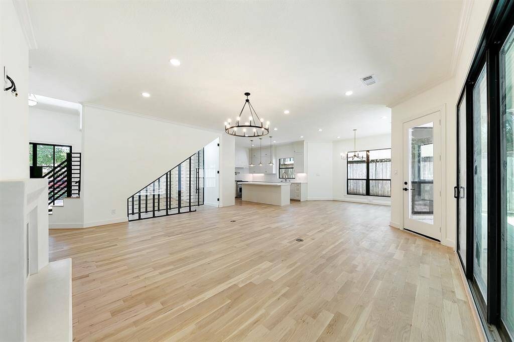 This spacious, open-concept living area features light wood flooring, large windows, and modern lighting. The kitchen has an island and sleek cabinetry, while sliding glass doors lead to the outdoor area, offering ample natural light.