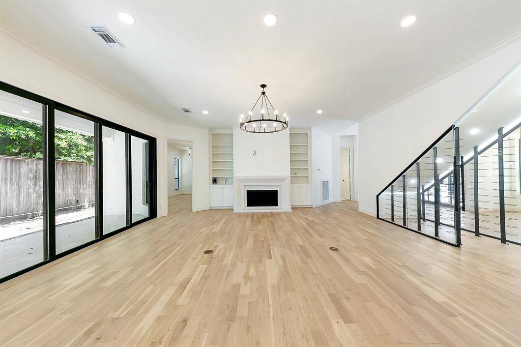 This spacious living area features hardwood floors, a modern chandelier, a sleek fireplace with built-in shelves, and large sliding glass doors leading to a patio. The open layout is complemented by a stylish staircase with glass railing.