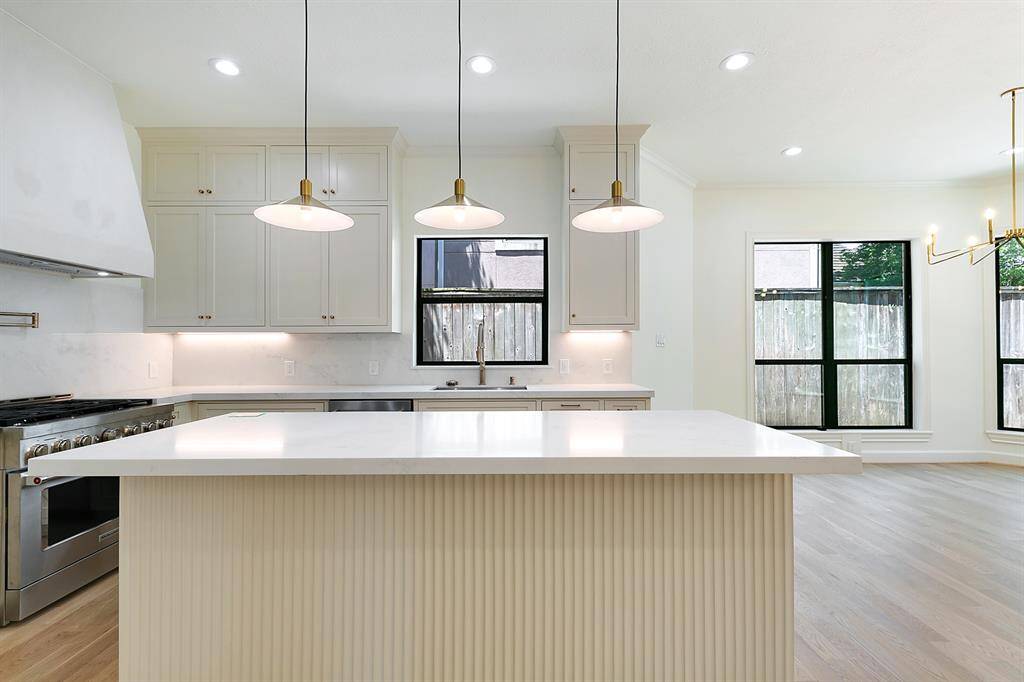 This modern kitchen features a spacious island with a sleek countertop, pendant lighting, a stainless steel range, and ample cabinetry. It offers abundant natural light from large windows, creating a bright and inviting space.