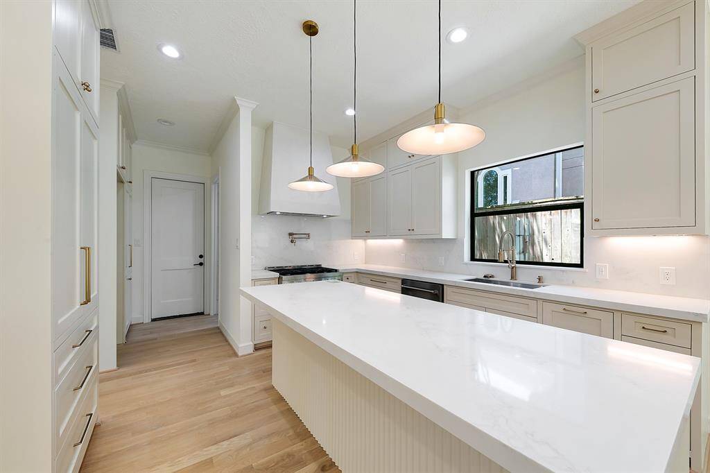 This modern kitchen features a spacious layout with light wood flooring, sleek white cabinetry, and a large island with a marble countertop. Pendant lights add elegance, while ample natural light streams through a nearby window.